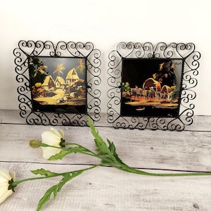 Peut inclure: Deux images encadrées en métal noir avec des volutes ornées. Les images présentent des scènes peintes d'un village et d'une caravane.