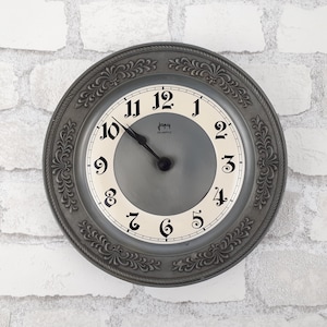 May include: A silver-toned wall clock with a white face and black Roman numeral hour markers. The clock has a decorative, ornate border and the text "Jam Quartz" is printed on the face.