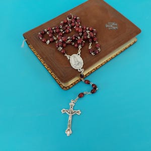 Puede incluir: Un rosario con cuentas rojo oscuro y un crucifijo plateado descansa sobre un libro marrón. El rosario presenta una pieza central plateada con una imagen religiosa. El libro tiene bordes dorados y un fondo azul.