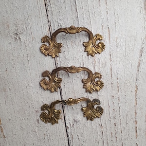 Peut inclure: Trois poignées de tiroir en laiton assorties présentant un design incurvé orné avec des volutes en forme de feuilles complexes à chaque extrémité. Chaque poignée possède deux petits trous de montage circulaires pour la fixation.