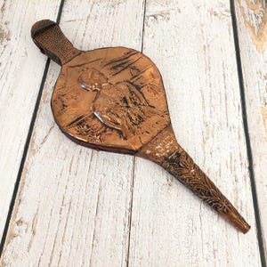 Può includere: Un mantice color rame con un design in rilievo di una figura e fogliame. L'impugnatura e l'ugello presentano motivi strutturati. Probabilmente utilizzato per un camino o una fucina. Articolo vintage.