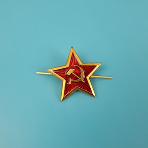 May include: A gold-colored star-shaped pin with a red center. The center features a gold hammer and sickle emblem. The pin has two horizontal bars extending from the star's points. The background is a solid light blue.
