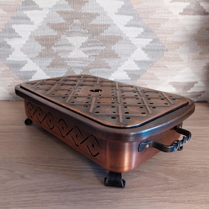May include: A copper-colored metal warming tray with a decorative patterned top and two black metal handles. The tray has four small feet and is designed to keep food warm.