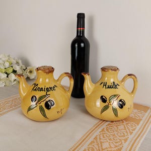 Peut inclure: Deux cruches en céramique jaune pour l'huile et le vinaigre avec des bouchons en liège. Les cruches ont la forme de théières avec des poignées des deux côtés. Les cruches sont décorées de branches d'olivier et d'olives noires. La cruche de gauche dit "Vinaigre" et la cruche de droite dit "Huile".