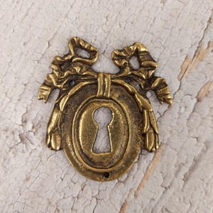 May include: Antique brass keyhole cover with an oval center and decorative bow and leaf accents. The keyhole is centered within the oval. The cover has a vintage aesthetic and is likely for a cabinet or drawer.