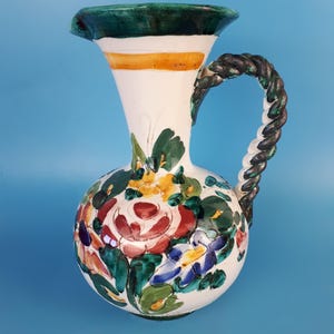 Peut inclure: Un pichet en céramique blanche avec un bord vert et jaune et une anse en corde torsadée. Le pichet est décoré de fleurs peintes à la main dans des tons de rouge, rose, bleu, jaune et vert.
