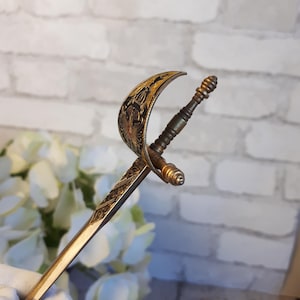 Peut inclure: Un ouvre-lettre ancien en forme de petite épée. La lame est dorée avec des motifs gravés complexes. La garde et la poignée sont également dorées, avec un design incurvé détaillé. L'ouvre-lettre est placé sur un fond flou.