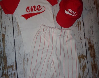 May include: A white and red baseball-themed outfit for a baby. The outfit includes a white shirt with red sleeves, red pants with white stripes, and a red baseball cap. The shirt has the word "one" written on it in red.
