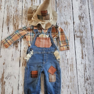 May include: A scarecrow costume consisting of a burlap hat with fabric patches, a long-sleeved plaid shirt in orange, blue, and tan, and blue denim overalls. The overalls feature a plaid chest pocket, straw accents, and various fabric patches.