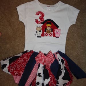 May include: A white t-shirt with a red barn, a cow, a pig, and the number 3 embroidered on it. The shirt is paired with a red, white, and blue patchwork skirt with a red and white checkered bow.