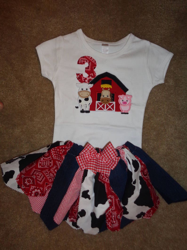 May include: A white t-shirt with a red barn, a cow, a pig, and the number 3 embroidered on it. The shirt is paired with a red, white, and blue patchwork skirt with a red and white checkered bow.