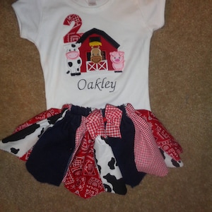 May include: White onesie with a red barn, cow, pig, and the number 2 embroidered on it. The onesie also has the name Oakley embroidered on it. The onesie is paired with a multi-colored skirt with red, white, and blue checkered fabric.
