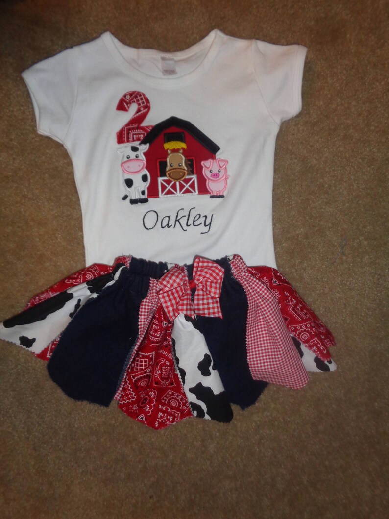 May include: White onesie with a red barn, cow, pig, and the number 2 embroidered on it. The onesie also has the name Oakley embroidered on it. The onesie is paired with a multi-colored skirt with red, white, and blue checkered fabric.