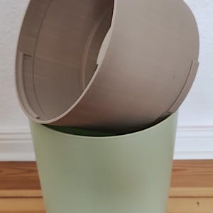 Op de afbeelding: Twee cilindrische bloempotten, de ene in de andere. De buitenste pot is lichtgroen van kleur en de binnenste pot is beige. De potten zijn gemaakt van een glad, mat materiaal. De foto is binnenshuis gemaakt.