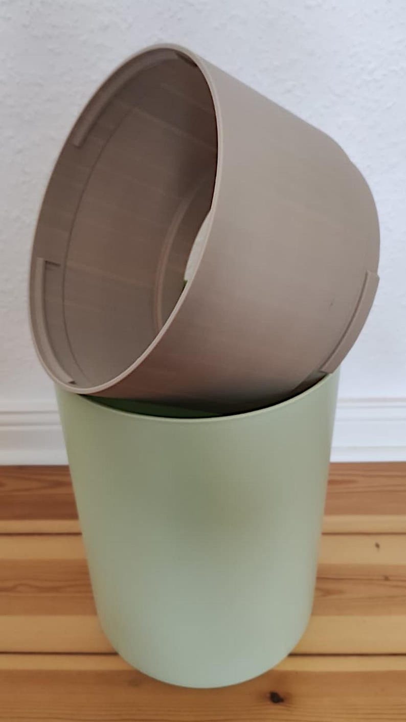 Op de afbeelding: Twee cilindrische bloempotten, de ene in de andere. De buitenste pot is lichtgroen van kleur en de binnenste pot is beige. De potten zijn gemaakt van een glad, mat materiaal. De foto is binnenshuis gemaakt.