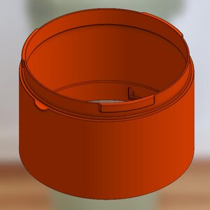 May include: An orange, cylindrical container with a rounded rim and a recessed handle. The container is set against a blurred, light green background, suggesting a product design or prototype.