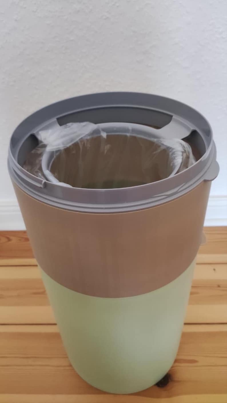 Op de afbeelding: Een tweekleurige afvalbak met een lichtgroen onderste gedeelte en een bruin bovenste gedeelte. De bak heeft een grijze rand en een doorzichtige plastic voering is zichtbaar. De bak staat op een houten vloer.