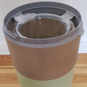 May include: A two-tone wastebasket with a light green lower section and a tan upper section. The rim is gray, and a clear plastic bag is visible inside. The wastebasket is on a wooden floor.