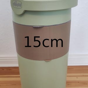 Op de afbeelding: Een lichtgroene luieremmer met een deksel en handvat. De emmer heeft een bruine band in het midden met de tekst "15cm". De emmer is ontworpen om gebruikte luiers in te bewaren en weg te gooien.