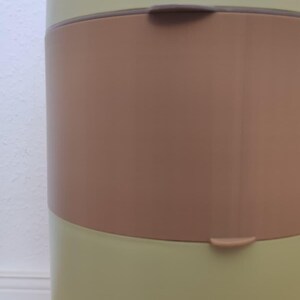 Op de afbeelding: Een lichtgroene en bruine luieremmer met een handvat en een bruine band in het midden. De emmer heeft een gebogen ontwerp en een deksel met de merknaam "chicco".