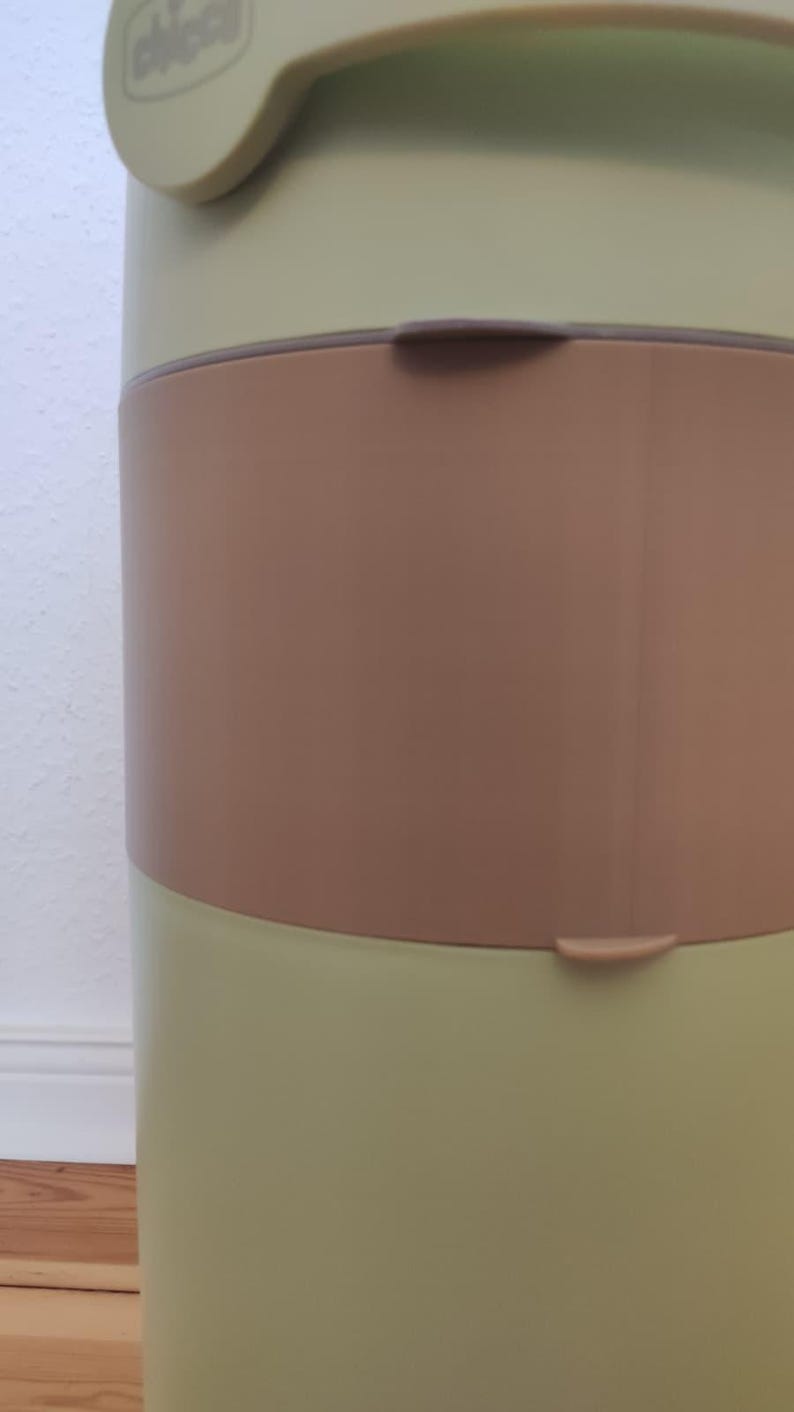 Op de afbeelding: Een lichtgroene en bruine luieremmer met een handvat en een bruine band in het midden. De emmer heeft een gebogen ontwerp en een deksel met de merknaam "chicco".