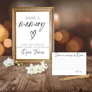 Puede incluir: Un letrero enmarcado con el texto "SHARE A memory" y un símbolo de corazón. Una tarjeta más pequeña dice "Share a memory of Ryan" con un bolígrafo. Flores blancas y una mesa de madera completan la escena.