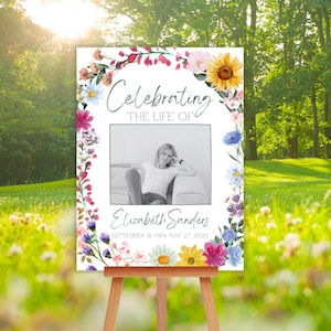 May include: A memorial sign on a wooden easel, featuring a photo of a woman and the text "Celebrating the Life of Elizabeth Sanders." The sign is framed with a floral arch of colourful flowers, including sunflowers and daisies. The dates of birth and death are also included.