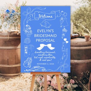 May include: A blue sign on an easel with white text and illustrations. The sign reads "Welcome" at the top, followed by "EVELYN'S BRIDESMAID PROPOSAL." Below, it says "Something borrowed, something blue, but most importantly, I need you!" and "NOVEMBER 2, 2026."
