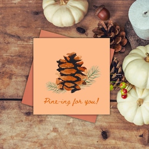 Op de afbeelding: Een vierkante wenskaart met een aquarel illustratie van een dennenappel en de tekst "Pine-ing for you!". De kaart staat op een perzikkleurige achtergrond, met een perzik envelop. De achtergrond bevat pompoenen en dennenappels.