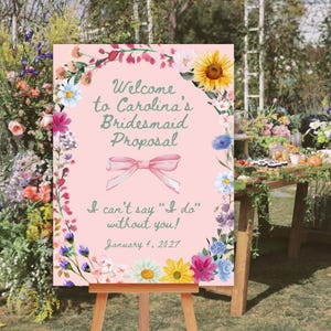 Könnte beinhalten: Ein rosa Schild auf einer Holzstaffelei, umrahmt von bunten Blumen. Auf dem Schild steht "Welcome to Carolina's Bridesmaid Proposal" mit einer Schleifenillustration und dem Text "I can't say 'I do' without you! January 4, 2027."