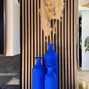 Peut inclure: Trois vases bleu cobalt, aux surfaces texturées, groupés, contiennent des herbes ornementales séchées de couleur beige. Les vases sont posés sur une surface noire, avec un mur à lattes en bois en arrière-plan. La scène est bien éclairée.