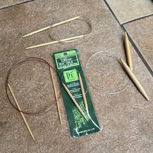 May include: A close-up of a package of Clover Takumi Bamboo Circular Knitting Needles, size 8 (5.00 mm), 16 inches (41 cm) long. The package is green and white with a picture of the needles on it. There are also several wooden knitting needles and a clear plastic cord in the image.