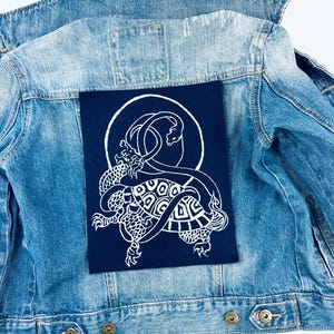 May include: A light blue denim jacket with a navy blue patch featuring a white illustration of a dragon-like creature within a circle. The jacket has a classic design with button closures and pockets.
