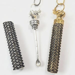 Könnte beinhalten: Zwei zylindrische Schnupflöffel mit Hirschkopf-Verzierungen und griechischem Mäander-Muster. Einer ist silberfarben mit passender Kette, der andere goldfarben mit passender Kette. Beide haben ein strukturiertes, sich wiederholendes Dreiecksmuster.