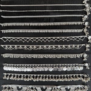 Può includere: Un set di dieci cavigliere d'argento con vari design, tra cui catene, campanelle e ciondoli a forma di moneta. Le cavigliere sono disposte in fila su una superficie di legno scuro.