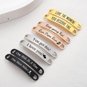 May include: An assortment of engraved metal shoe tags in gold, rose gold, silver, and black. The tags feature motivational phrases such as "Love the Moment" and "One Mile At A Time". These tags are designed for runners and fitness enthusiasts.
