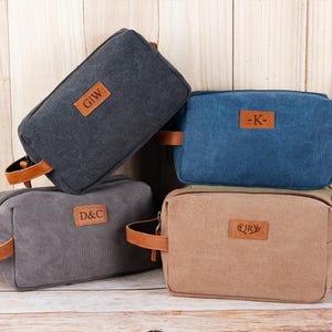 Könnte beinhalten: Vier Kulturbeutel aus Canvas in Grau-, Blau- und Beigetönen, jeweils mit einem braunen Ledergriff und einem rechteckigen Lederaufnäher mit Initialen. Im Hintergrund ist eine Weinflasche zu sehen.