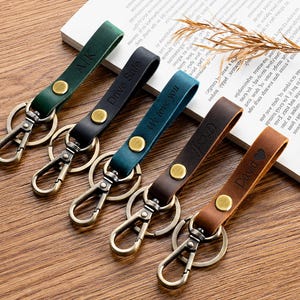 May include: Five leather keychains in green, black, teal, dark brown, and tan, each featuring a brass-colored snap button, a metal lobster clasp, and a split ring. The straps are embossed with text including AK, Drive Safe, We love you, and David.