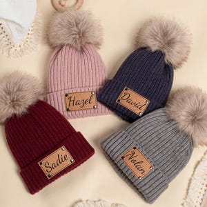 Puede incluir: Cuatro gorros de punto personalizados con pompones de piel sintética. Los gorros son de color burdeos, rosa, azul marino y gris. Cada gorro tiene un parche de cuero con un nombre: "Sadie", "Hazel", "David" y "Nolan".
