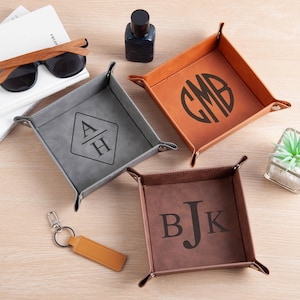 May include: Four personalized leather valet trays in gray, brown, and tan. Each tray features initials or a monogram. Sunglasses, a small bottle, a keychain, and a small plant are also visible.
