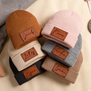 May include: A selection of personalised beanies in shades of brown, pink, grey, and beige. Each beanie has a leather patch with a name in a script font. The names visible are Jacob, Nova, Lily, Aiden, Ezra, and Isaac.