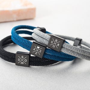 Op de afbeelding: Drie gepersonaliseerde armbanden in zwart, blauw en grijs. Elke armband heeft een geweven touwontwerp en een zwarte vierkante sluiting met gegraveerde initialen en cijfers. De armbanden worden gepresenteerd op een lichtgrijze ondergrond.