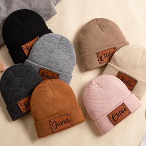 Peut inclure: Plusieurs bonnets personnalisés de couleurs variées, dont noir, gris, marron et rose. Chaque bonnet est orné d'un écusson en cuir avec un prénom, comme "Stella" ou "Chloe". Les bonnets sont disposés sur une surface neutre.