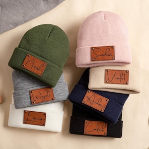 May include: A collection of personalized beanies in various colors, including green, pink, gray, white, navy, and black. Each beanie features a brown leather patch with a name in a stylish script font. The names include Noah, Sadie, Wyatt, Violet, Liyma, Aiden, and Caleb.