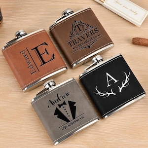 May include: Four personalized leather-wrapped hip flasks with different designs. The flasks are brown, black, and gray. The designs include an initial, a crest, antlers, and a tuxedo. The text on the flasks reads "Travers - Groomsman", "Andrew - Groomsman", "E. Piarpet", and "A".