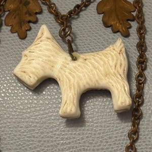May include: A vintage necklace featuring a cream-colored terrier pendant. The dog charm is attached to a bronze-toned chain with decorative leaf accents. The necklace has a classic, antique aesthetic.