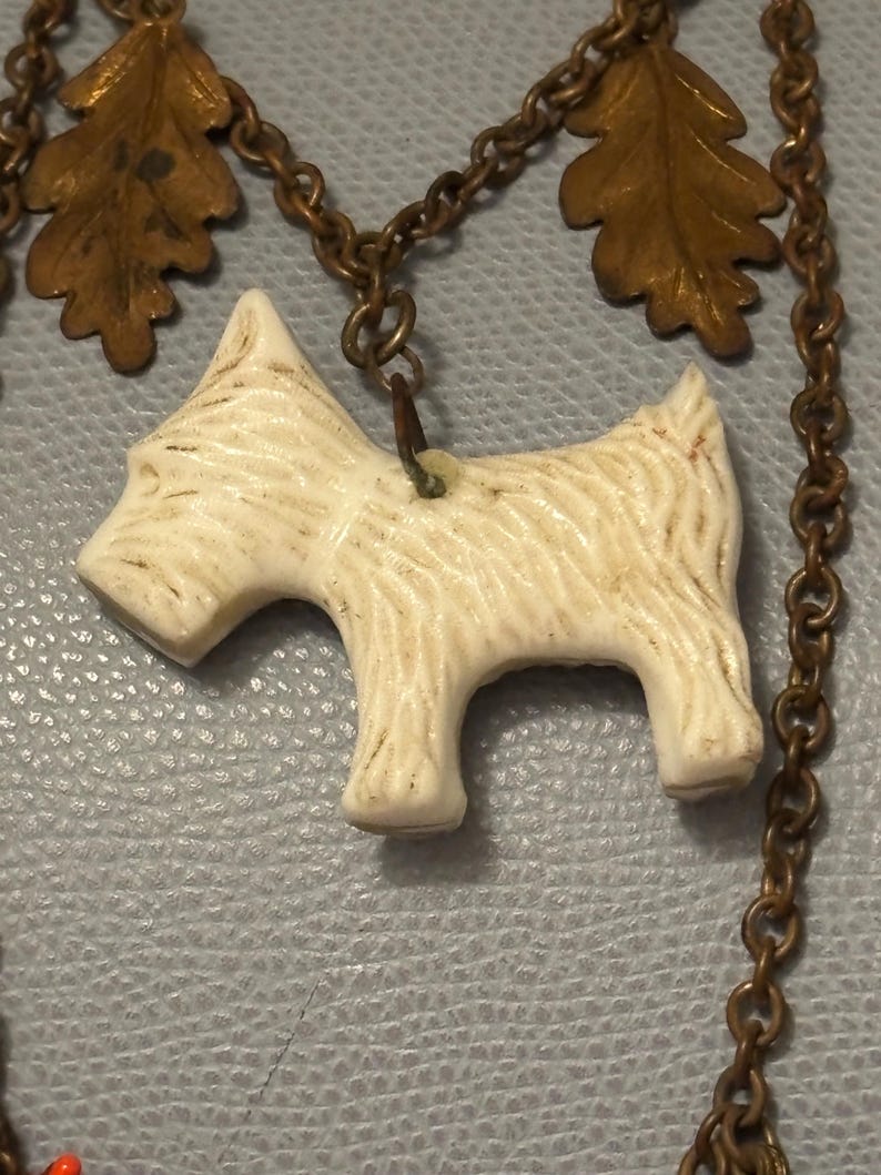 May include: A vintage necklace featuring a cream-colored terrier pendant. The dog charm is attached to a bronze-toned chain with decorative leaf accents. The necklace has a classic, antique aesthetic.