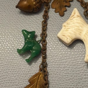 May include: A vintage necklace featuring woodland-themed charms. The charms include a green cat, a brown acorn, a white dog, and orange and bronze-colored leaves. The charms are connected by a bronze-colored chain.