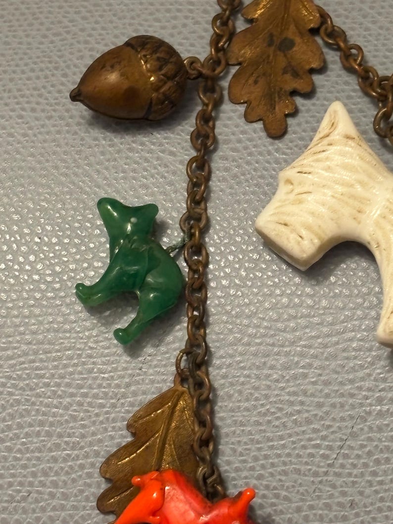 May include: A vintage necklace featuring woodland-themed charms. The charms include a green cat, a brown acorn, a white dog, and orange and bronze-colored leaves. The charms are connected by a bronze-colored chain.