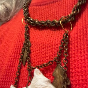 May include: A vintage necklace with a bronze chain and decorative charms. The charms include a white dog figurine, a leaf, and a small acorn. The necklace is worn over a red sweater.